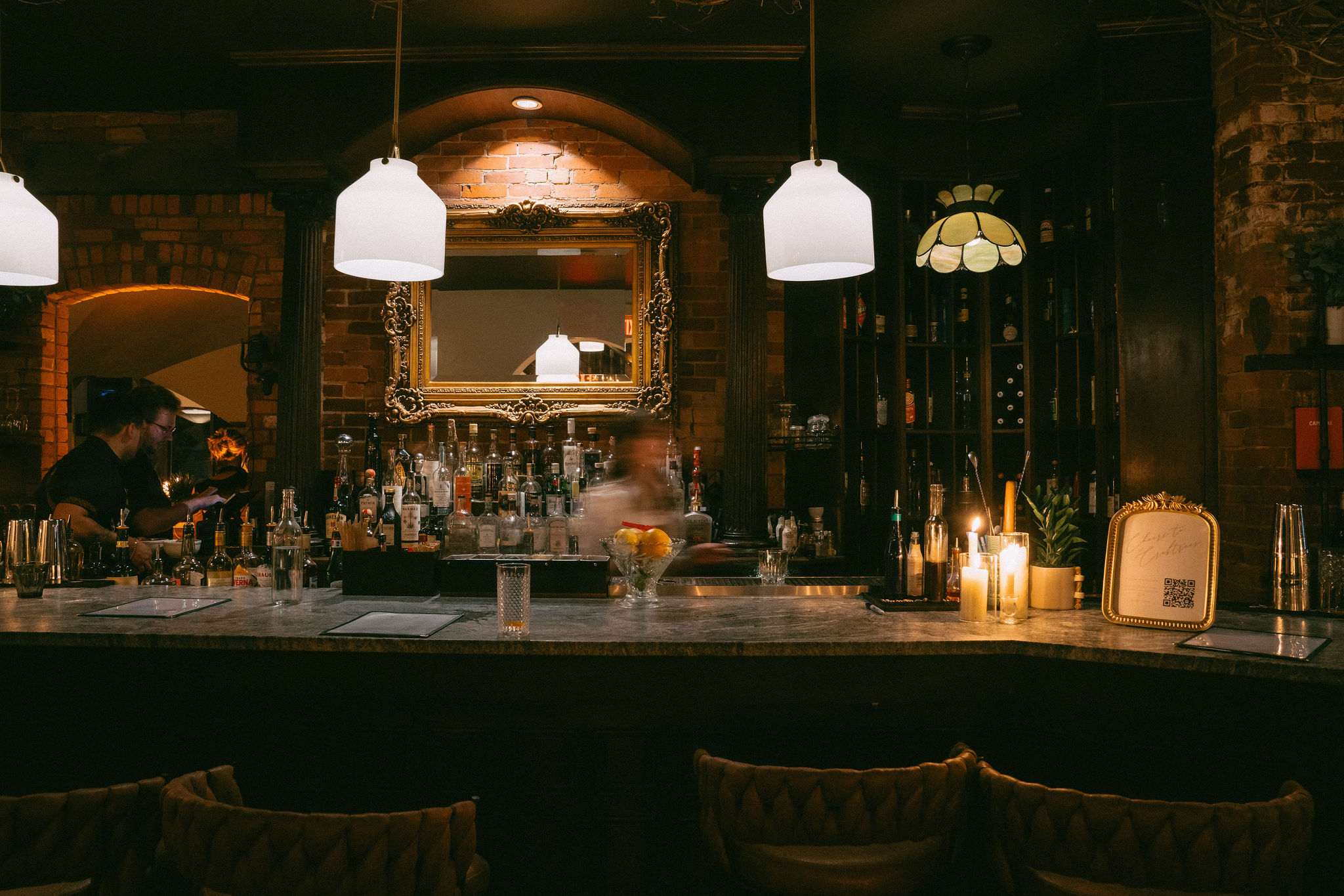 The Alexander bar in the evening — bartenders at work, candlelight, exposed brick, and ornate mirror